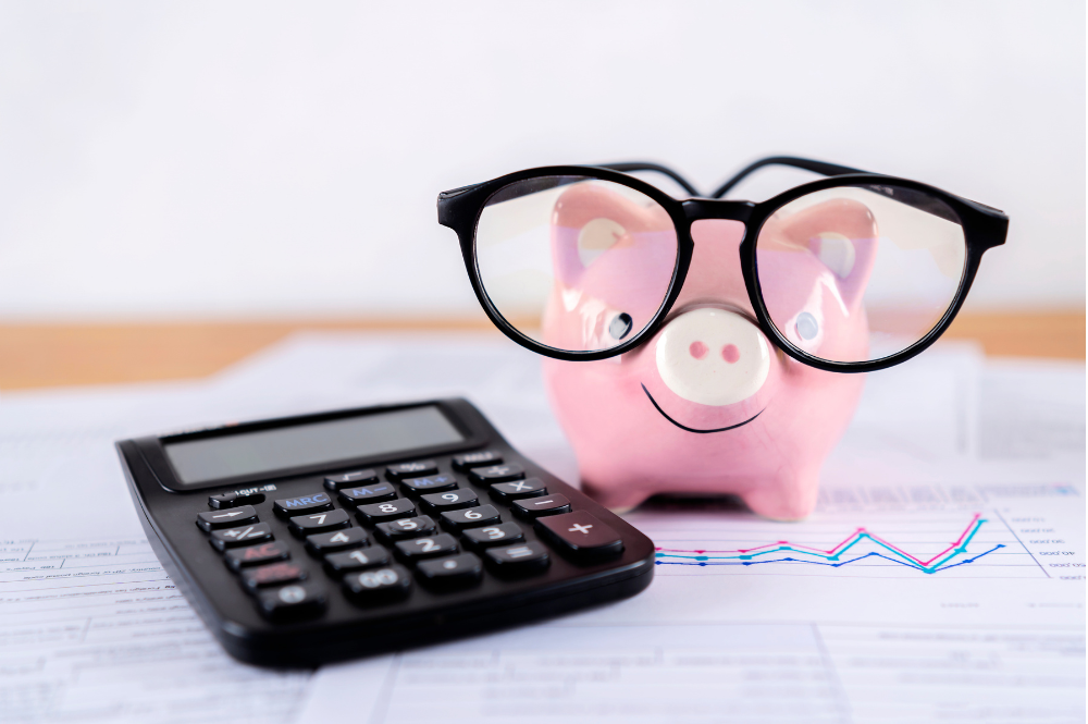 A piggybank with glasses next to a calculator and paper with a graph on it, illustrating how to build an emergency fund.